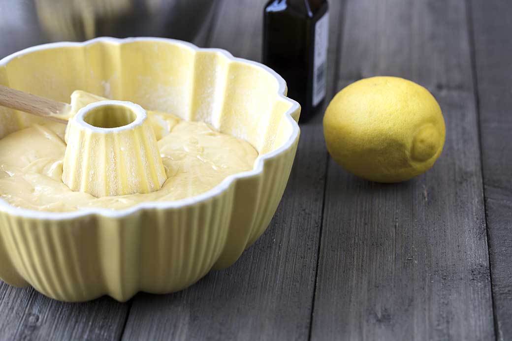 A yellow Bundt pan sits full of batter next to a bottle of vanilla and a lemon on a wooden surface.
