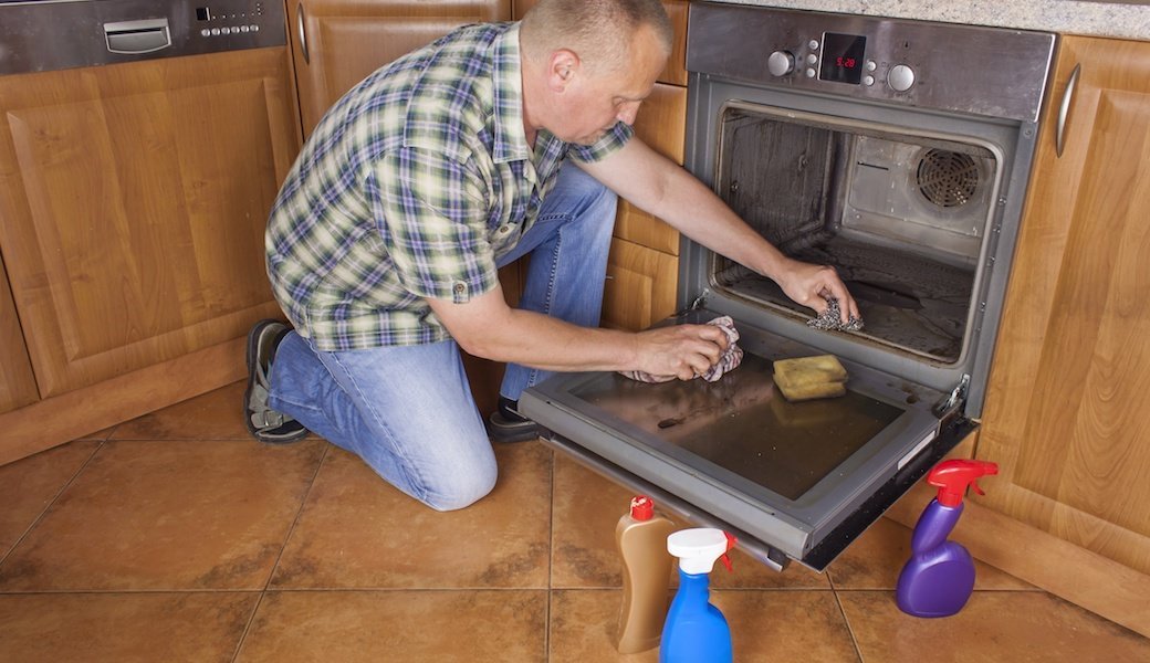 A man kneels next to an open oven with a variety cleaning bottles next to him as he cleans.