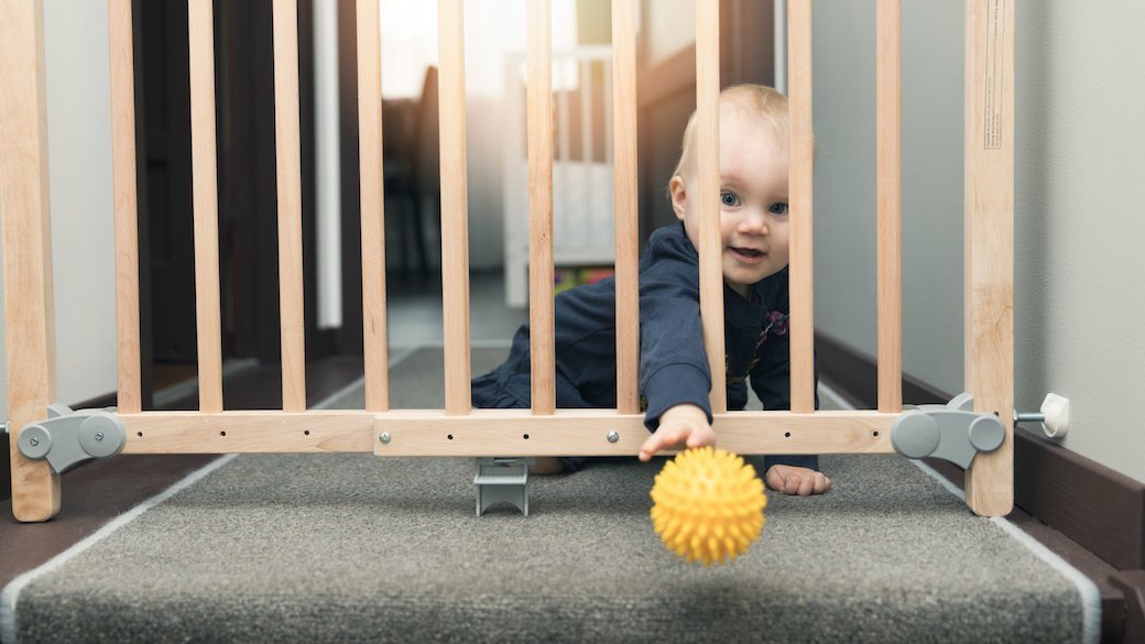 A baby reaches their arm through a baby gate at the top of the stairs in an attempt to reach a yellow ball.