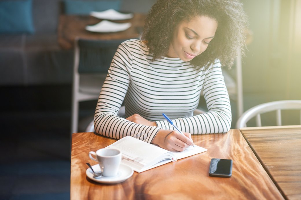A young woman with curly hair is wearing a black and white striped shirt and sitting at a wooden table writing in a journal.