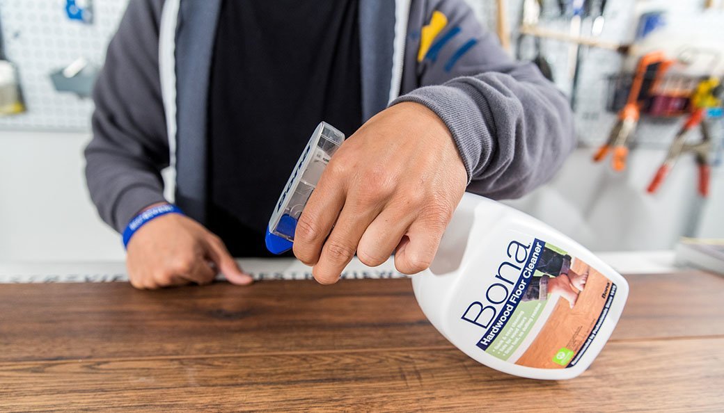 A person wearing a black shirt and gray cardigan uses a spray on hardwood floor cleaner to clean a wood surface.