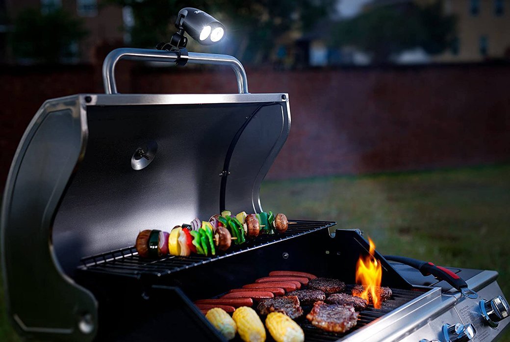A grill light illuminates a propane grill with kebabs, potatoes, hamburgers, and hot dogs on it.
